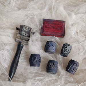 Pode incluir: Um conjunto de ferramentas de impressão vintage, incluindo um carimbo de rolo com um design floral, vários pequenos carimbos cilíndricos com padrões ornamentados e uma caixa vermelha com texto. As ferramentas estão dispostas sobre uma superfície branca texturizada.