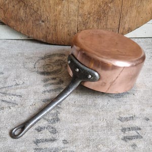 Puede incluir: Una cacerola de cobre vintage con un mango de hierro negro. La cacerola tiene un fondo redondeado y un borde acampanado. El mango está unido al lado de la cacerola con remaches.