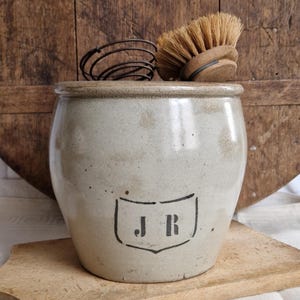 Può includere: Un vaso in gres vintage con coperchio, con un disegno a scudo nero con le lettere "J R". Una frusta e una spazzola di legno sono appoggiate sopra. Il vaso è su un tagliere di legno, con uno sfondo di legno rustico.
