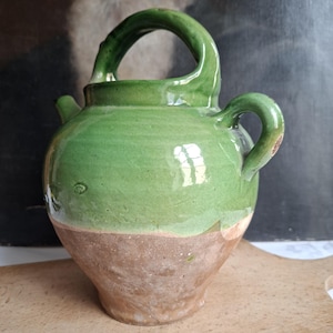 Peut inclure: Une cruche en céramique avec un corps arrondi et une base étroite. La partie supérieure présente une glaçure verte brillante, tandis que la section inférieure reste non émaillée, révélant une texture d'argile brune naturelle.