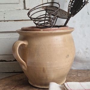 May include: A large, beige ceramic crock with a single handle. The crock is filled with two vintage metal kitchen utensils, a whisk and a slotted spoon.
