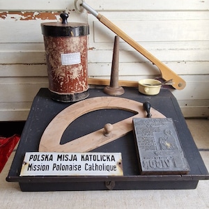 Peut inclure: Une boîte en bois noire avec un couvercle à charnières, contenant un rapporteur en bois, un petit récipient en métal, un cône en bois, un petit bol en métal et un bloc d'impression en métal avec le texte "Vigor" et "Blauw & Kuyt" en blanc. La boîte a également une pancarte blanche avec le texte "Polska Misja Katolicka Mission Polonaise Catholique".