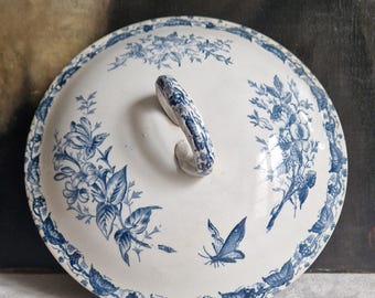 Puede incluir: Tapa de cerámica blanca con diseños florales y mariposas azules. La tapa tiene un asa curva y un borde decorativo. Ideal para la decoración de la cocina o el comedor.