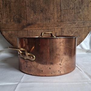 Op de afbeelding: Een ronde koperen pan met aan weerszijden een gepolijst messing handvat en een bijpassend messing handvat op het deksel. De buitenkant van de pan heeft een warme, metalen koperen afwerking met subtiele gebruikssporen en natuurlijke patina.