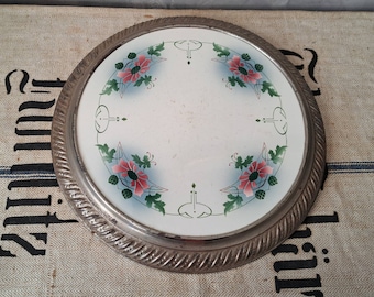 May include: A vintage silver-plated cake stand with a white ceramic top featuring a floral design with pink and green flowers. The stand has a decorative, raised edge.