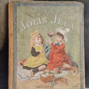 May include: Vintage children's book titled "Mes Jolis Jeux" featuring illustrations of two children playing. The cover shows a girl in a yellow dress and a boy in a red dress. The book is published by HACHETTE.