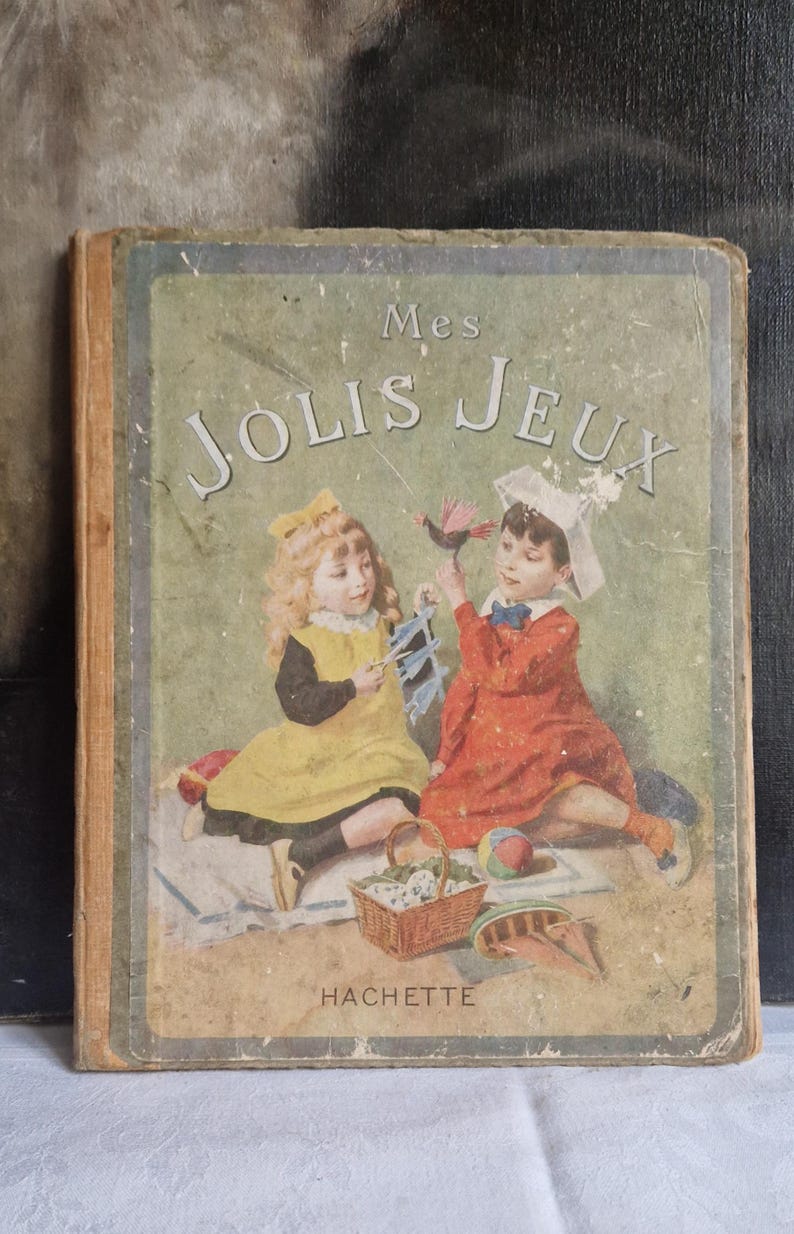 May include: Vintage children's book titled "Mes Jolis Jeux" featuring illustrations of two children playing. The cover shows a girl in a yellow dress and a boy in a red dress. The book is published by HACHETTE.