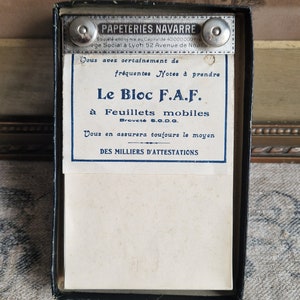 Può includere: Una scatola vintage nera con un'etichetta bianca che recita "Le Bloc F.A.F. à Feuillets mobiles Broveté S.G.D.G. Vous en assurera toujours le moyen DES MILLIERS D'ATTESTATIONS". L'etichetta è circondata da un bordo blu. La scatola è chiusa e ha due chiusure in metallo argentato.