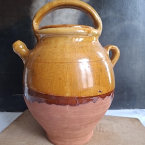 Pode incluir: Um jarro de cerâmica vidrada com corpo arredondado, apresentando uma secção superior em amarelo-mostarda brilhante e uma base em terracota mate. O recipiente inclui uma pega arqueada proeminente no topo, um pequeno bico e uma segunda pega pequena.