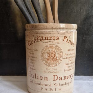 Peut inclure: Pot en céramique ancien avec l'inscription "Confitures Fines" et "Julien Damoy PARIS" en lettres marron. Il contient des ustensiles en bois. Le pot est blanc cassé avec une finition vieillie, suggérant son âge.