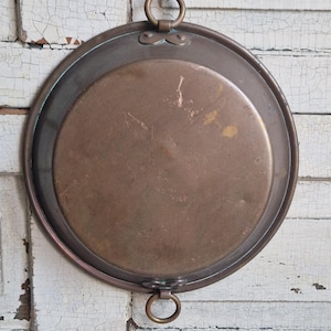 Puede incluir: Una bandeja redonda de cobre antiguo con borde elevado y dos asas de anillo. La bandeja tiene una pátina oscura y envejecida y está colocada sobre un fondo de madera blanca desgastada. El diámetro de la bandeja es de unos 30 cm.