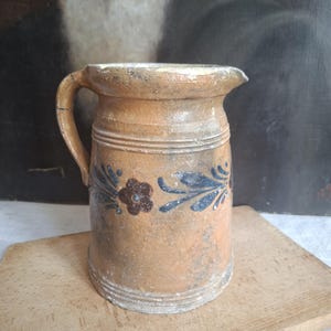 Könnte beinhalten: Ein zylindrischer Krug aus beigefarbenem Steingut mit einem einzelnen Henkel und einem Ausguss. Die Außenfläche zeigt ein handgemaltes Blumenmotiv in dunkelblauen und braunen Tönen sowie horizontale Rillenbänder am Hals und am Boden des Gefäßes.