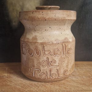 Könnte beinhalten: Beige Keramiktopf mit Deckel und der Prägung „Poubelle de Table“. Der Topf hat eine gesprenkelte Oberfläche und eine leicht ausgestellte Form. Ein kleiner, runder Deckel sitzt oben auf. Der Topf ist wahrscheinlich für die Küche gedacht.