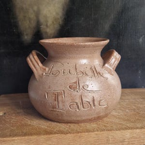 Könnte beinhalten: Eine beige Keramikvase mit zwei Henkeln und den Worten "Bubulle de Table" in die Oberfläche geätzt. Die Vase hat einen runden Körper und einen ausgestellten Rand. Sie steht auf einem Holzschneidebrett.