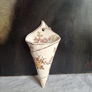 Puede incluir: Un florero de pared de cerámica color crema con forma de cono y parte superior puntiaguda. La superficie presenta un delicado patrón floral con flores amarillas, rosas y azules sobre tallos marrones. Un pequeño orificio superior permite colgarlo.