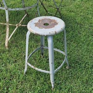 May include: A gray metal stool with a round seat and a single ring footrest. The stool has a worn, rustic look with signs of rust.