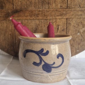 May include: A beige stoneware crock with a blue floral design holds two burgundy candles. The crock sits on a white surface, with a wooden background. The rustic design is suitable for home decor.