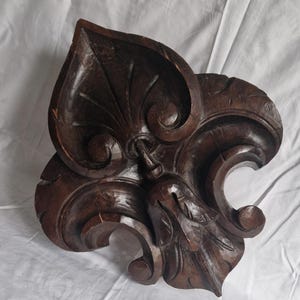 Puede incluir: Elemento decorativo de madera tallada, de color marrón oscuro, con un diseño de hoja y volutas estilizadas. Los detalles incluyen vetas talladas en la hoja y un elemento en forma de campana. Probablemente sea un detalle arquitectónico decorativo.