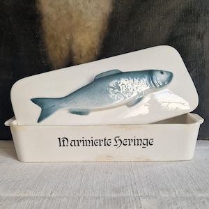May include: A rectangular, off-white ceramic dish with a lid. The lid features a raised, light blue fish design. The dish is labeled "Marinierte Heringe" in black script. A vintage kitchenware item.
