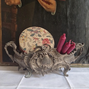 Op de afbeelding: Een sierlijke, zilverkleurige kandelaar met drie magenta kaarsen. Een ronde doos met een bloemenpatroon staat achter de kaarsen. De kandelaar heeft een decoratief, krullend ontwerp en staat op een wit oppervlak.