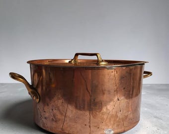 Op de afbeelding: Een ronde koperen kookpan met bijpassend deksel en twee handgrepen aan de zijkanten. Het buitenoppervlak van de pan vertoont een warme, metalen koperkleur met zichtbare gebruikssporen, waaronder kleine vlekjes en gepolijste handgrepen.