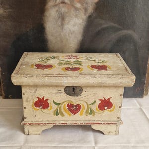 May include: A small, antique wooden box with a hinged lid. The box is painted cream with red and yellow heart and floral designs. It has a metal lock and sits on small feet. The box is likely used for storing small items.