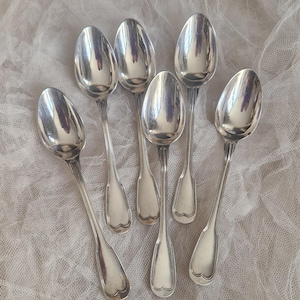 Op de afbeelding: Zes zilveren lepels gerangschikt op een witte, getextureerde stof. De lepels hebben een klassiek ontwerp met lange stelen en hartvormige bakken. De lepels zijn zeer reflecterend.