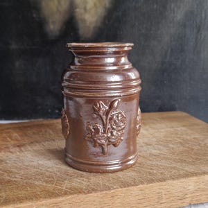 Könnte beinhalten: Ein brauner Keramiktopf mit floralem Reliefdesign. Der Topf hat eine zylindrische Form mit leicht ausgestelltem Oberteil und steht auf einer Holzoberfläche. Das florale Design wiederholt sich um den Topf.