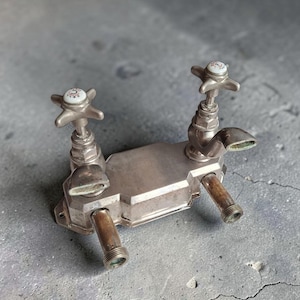 Könnte beinhalten: Eine Vintage-Armatur aus Metall mit zwei kreuzförmigen Griffen und weißen Keramikeinsätzen, die mit C und H gekennzeichnet sind. Das Objekt hat einen rechteckigen Mittelkörper, zwei gebogene Ausläufe und zwei nach unten gerichtete Kupferrohre.