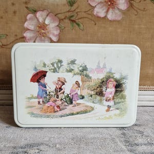 Può includere: Scatola vintage in metallo bianco con un'illustrazione colorata di quattro bambini che giocano in un giardino. I bambini indossano abiti colorati e tengono in mano attrezzi da giardinaggio e un ombrello. La scatola di metallo ha una forma rettangolare con un bordo leggermente rialzato.