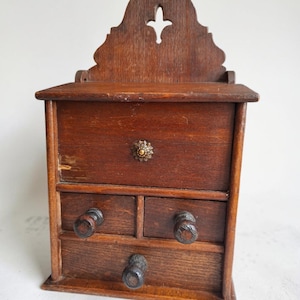 May include: A dark brown wooden wall-mounted storage unit featuring a tall backboard with a cutout design, a large top compartment with a decorative metal knob, and three smaller lower drawers each equipped with a round wooden pull handle.