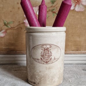 May include: A white ceramic jar with a brown design of a ship and anchor. The jar has three pink candles inside.