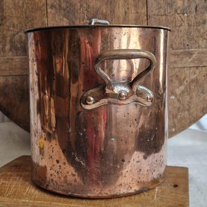 May include: An antique copper cooking pot with a lid and two handles. The cylindrical pot sits on a wooden chopping board. The copper has an aged patina, showing signs of use. A classic piece of kitchenware, perfect for traditional cooking.