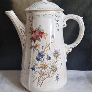 Könnte beinhalten: Antike weiße Keramik-Kaffeekanne mit Deckel, verziert mit Blumenmustern in Rot, Blau und Gelb. Die Kanne hat einen gebogenen Ausguss, einen dekorativen Griff und goldfarbene Verzierungen. Ein Vintage-Stück.