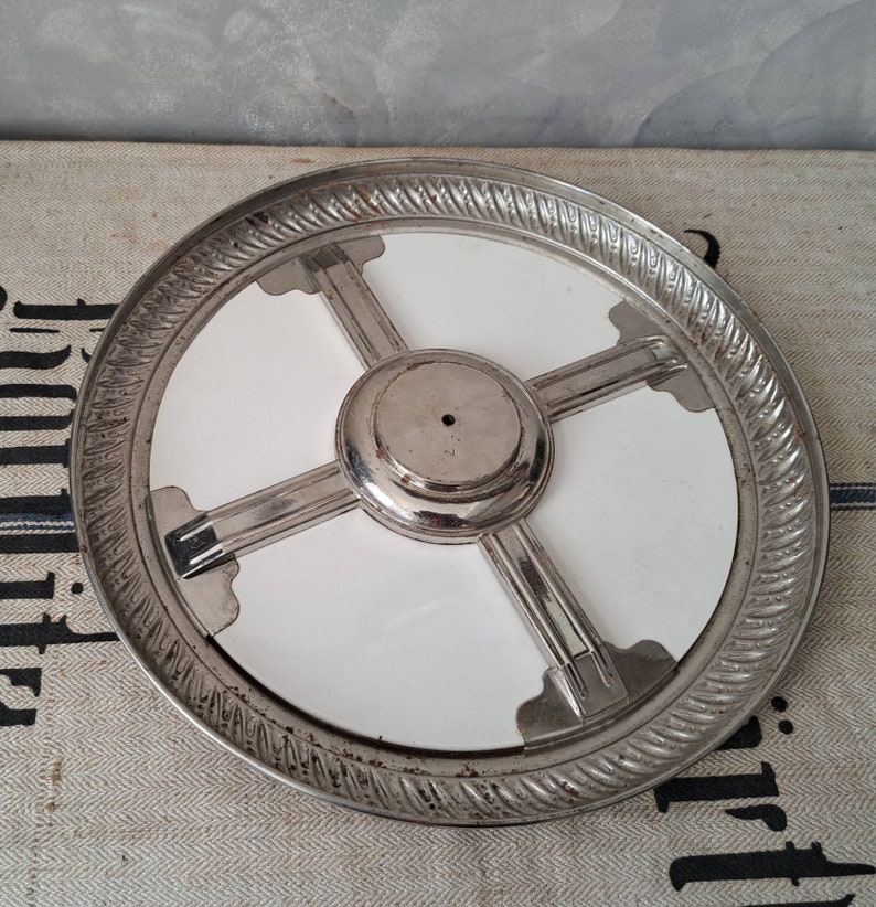 Op de afbeelding: Een zilveren metalen taartstandaard met een wit keramisch midden en een decoratieve touwachtige rand. De standaard heeft vier metalen armen die zich uitstrekken van het midden tot aan de rand van de standaard.