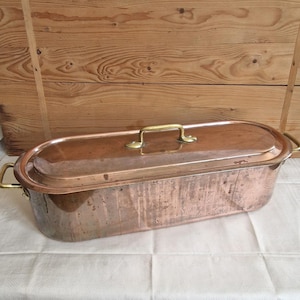 Pode incluir: Uma panela de cozinha oval de cobre com tampa e alças de latão. A panela tem formato retangular com extremidades arredondadas. A tampa tem uma única alça no centro. O exterior da panela mostra sinais de uso e idade.