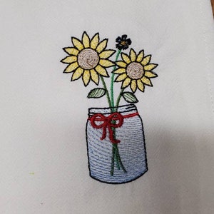 May include: Embroidered kitchen towel with a design of a mason jar filled with sunflowers and a red bow.