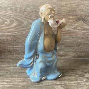 May include: A ceramic figurine of a man wearing a blue robe and holding a small red and white object in his hand. The figurine has a white beard and a brown face.