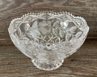 May include: A clear glass bowl with a cut glass design featuring floral motifs. The bowl has a pedestal base.