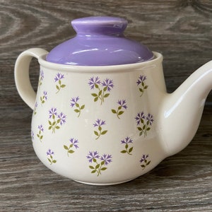 Könnte beinhalten: Eine weiße Teekanne mit einem lila Deckel und einem Blumenmuster aus kleinen lila Blüten und grünen Blättern.