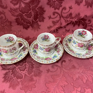 Puede incluir: Tres tazas de té blancas con motivos florales y ribetes dorados sobre platillos blancos con motivos florales a juego. Los platillos tienen un borde dorado.