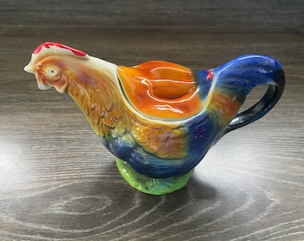 May include: A ceramic teapot shaped like a rooster with a blue, orange, brown and green glaze. The teapot has a lid and a handle.