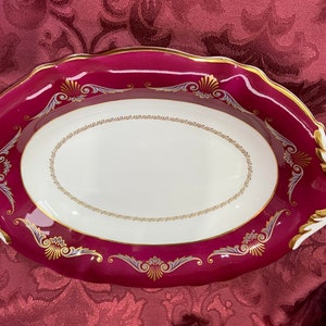 May include: An oval serving dish with a deep crimson red rim and a white centre. The rim is decorated with a gold trim and a delicate floral pattern in gold and silver.
