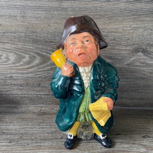 Puede incluir: Una figurita de cerámica de un hombre con un abrigo verde, un sombrero marrón y sosteniendo un cuerno amarillo. Está sentado en una roca y sosteniendo un rollo en su otra mano.