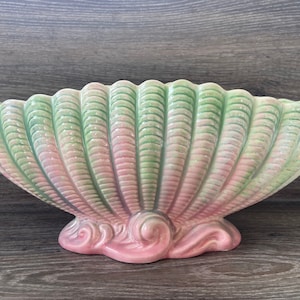 Puede incluir: Una jardinera de cerámica con forma de concha en tonos rosa y verde. La jardinera tiene un borde festoneado y una superficie texturizada, con una base decorativa con detalles en espiral. La jardinera está diseñada para parecerse a una gran concha.