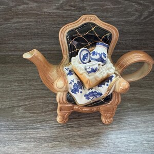 May include: A novelty teapot in the shape of a chair, with a brown and black frame. The teapot has a blue and white floral pattern, with a miniature teacup, saucer, and jug on top. A unique piece of kitchenware.