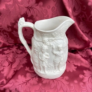 Puede incluir: Jarra de cerámica blanca con un diseño en relieve de dos figuras que sostienen uvas y vides. La jarra tiene un asa con un diseño de hoja.