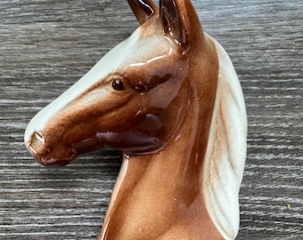 Puede incluir: Una figura de cerámica con forma de cabeza de caballo. La cabeza del caballo es marrón y blanca con un acabado brillante. La figura es un artículo decorativo, probablemente para exhibición.