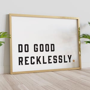 Puede incluir: Un cuadro enmarcado con las palabras "DO GOOD RECKLESSLY" en letras negras en negrita sobre un fondo blanco. El marco es dorado y hay una pequeña estrella dorada al final del texto. El cuadro está apoyado contra una pared.