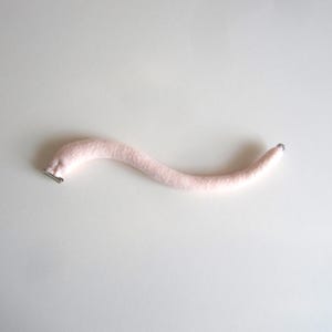 May include: A light pink, curved, plush tail with a silver metal clip at one end. The tail is made of a soft, fuzzy material and has a slight curve. The tail is a solid colour and is laying on a white surface.